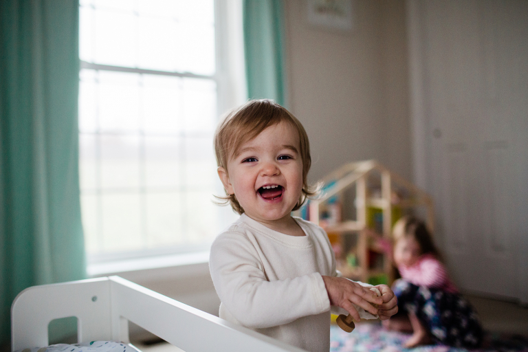 little girl laughing and smiling at the camera with big sister in the background playing with a dollhouse