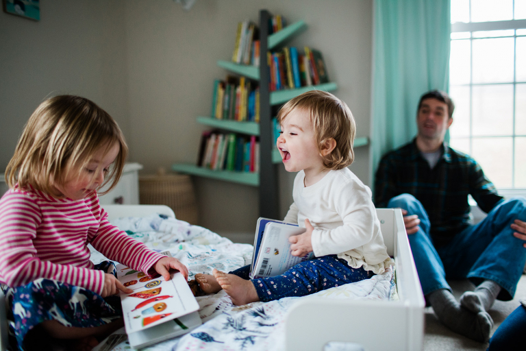 documentary photograph of two girls sitting on a bed reading to each other with dad in background