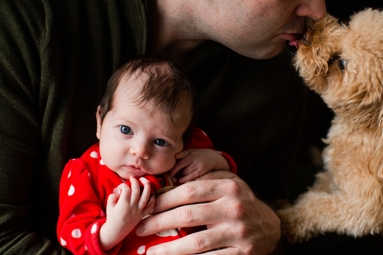 Lifestyle photo of newborn girl with puppy kissing her dad in the background
