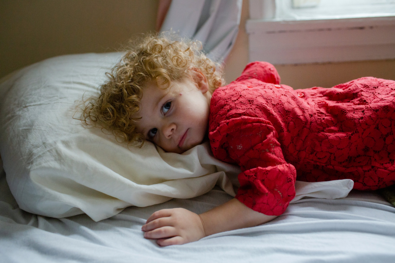 Documentary Photograph of toddler girl in red dress laying on a bed and gazing at camera