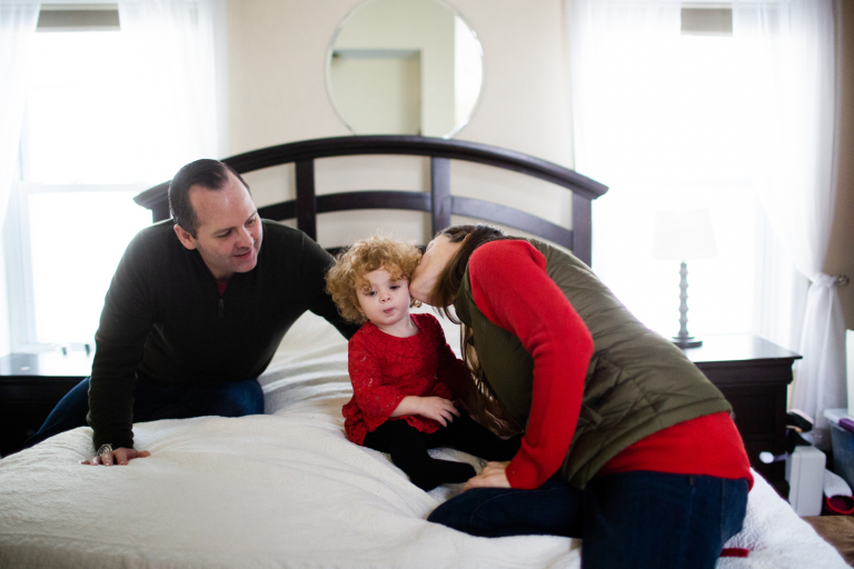 Image of mom and dad kissing toddler girl sitting on their bed