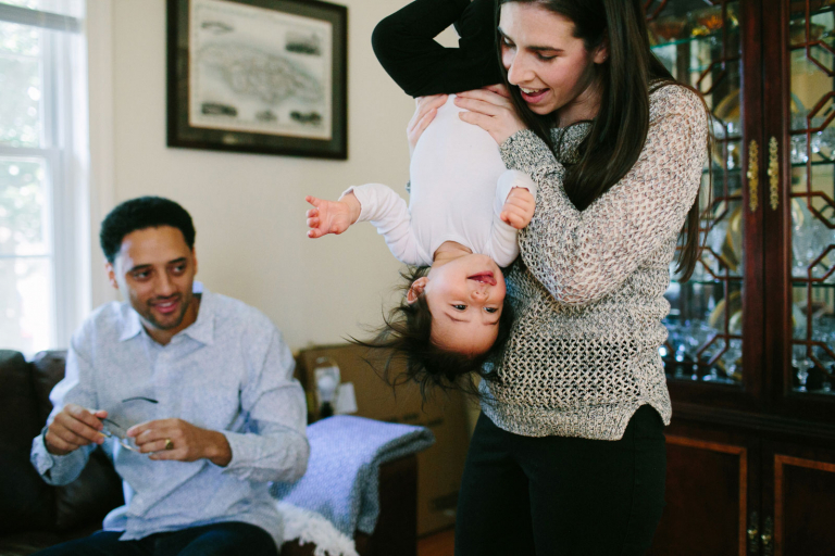Image of mom holding toddler girl upside down while dad smiles in the background