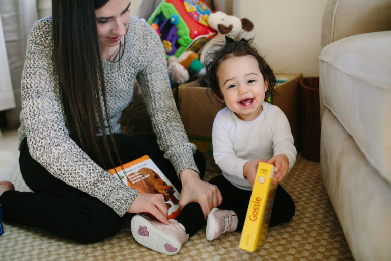 Documentary image of toddler girl looking at books and smiling at camera while her mom puts her shoes on