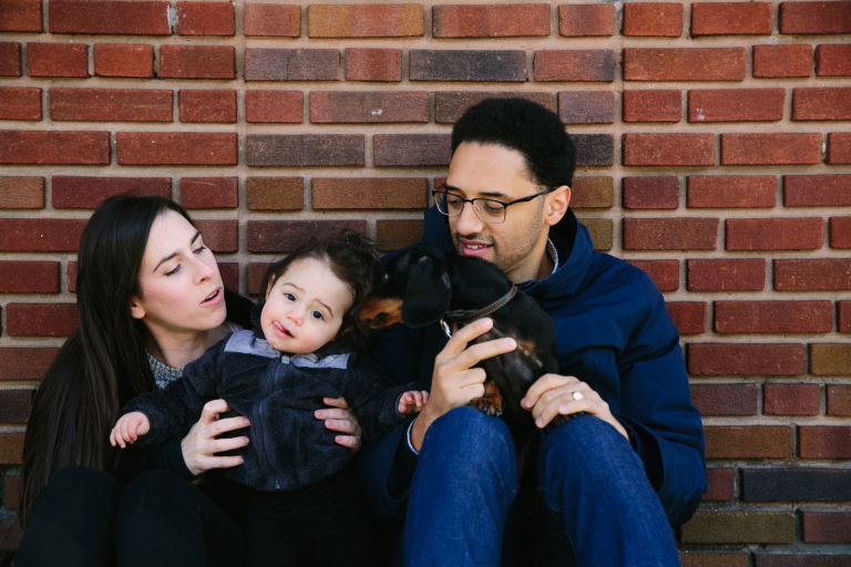 Lifestyle image of a family in front of a brick wall with dachshund kissing toddler girl