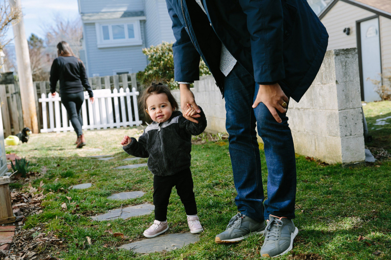 Image of dad walking, holding toddler girl's hand with mom in the background