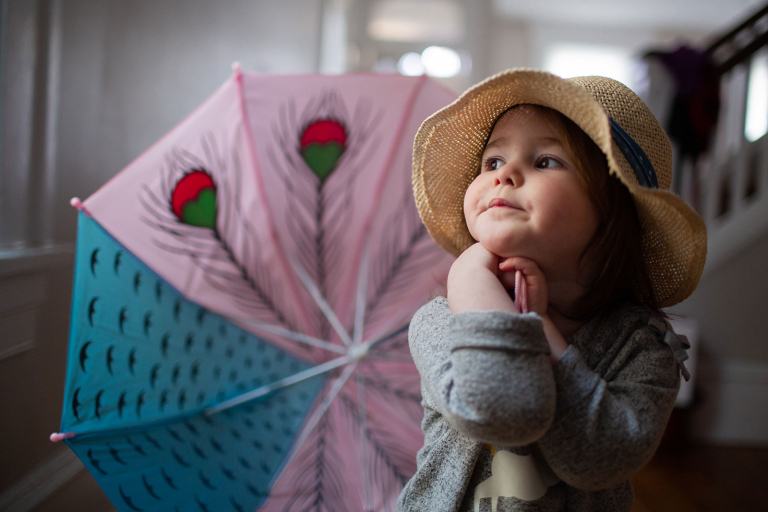 Little girl with straw hat, holding umbrella and looking out the window.