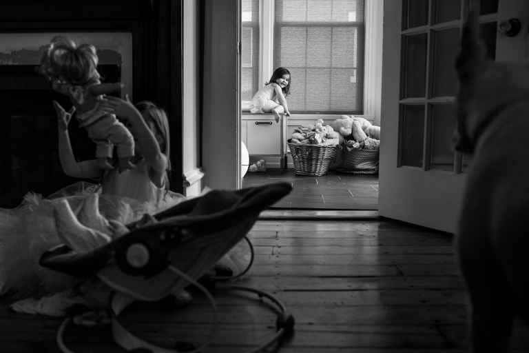 Black and white photo of toddler girl climbing on cabinet in the background with baby, older sister tossing doll and dog in the foreground.