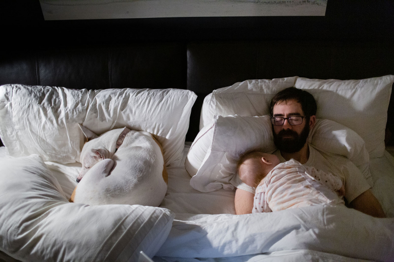 Documentary photo of baby sleeping on her dad's chest with white dog sleeping on the bed next to them.