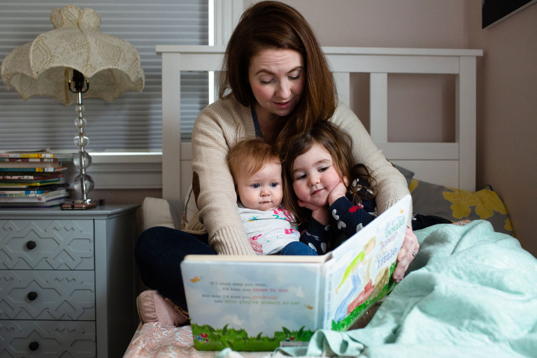 Documentary photo of mom reading books to baby and toddler girl on bed.