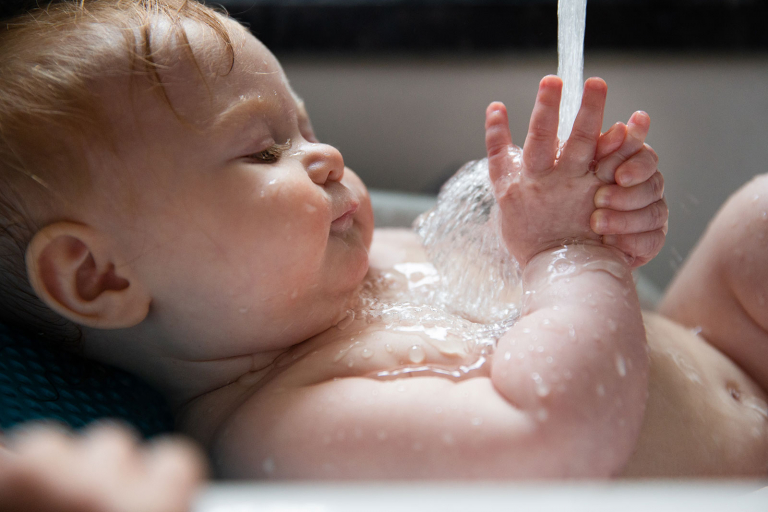 Documentary photo of baby being bathed in the sink, looking at the water running into her hands. 
