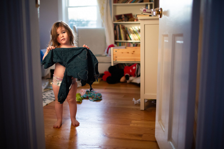 Toddler girl standing in just a diaper while holding her clothes in front of her.