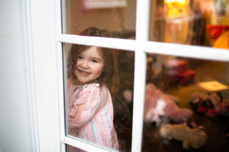 Toddler girl looking out through window with reflection of dog in the window. 