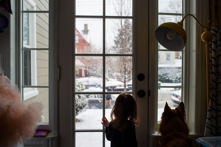 Conshohocken snowy day toddler girl and dog looking out the window at dad cleaning off car.