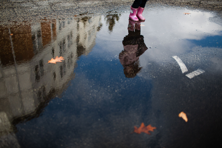 Reflection of little girl in puddle with pink boots and blue sky.