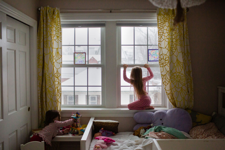 Little girl sitting on windowsill looking out at snowy scene with toddler girl playing legos