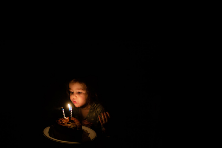 Image of little girl blowing out three candles surrounded by blackness.