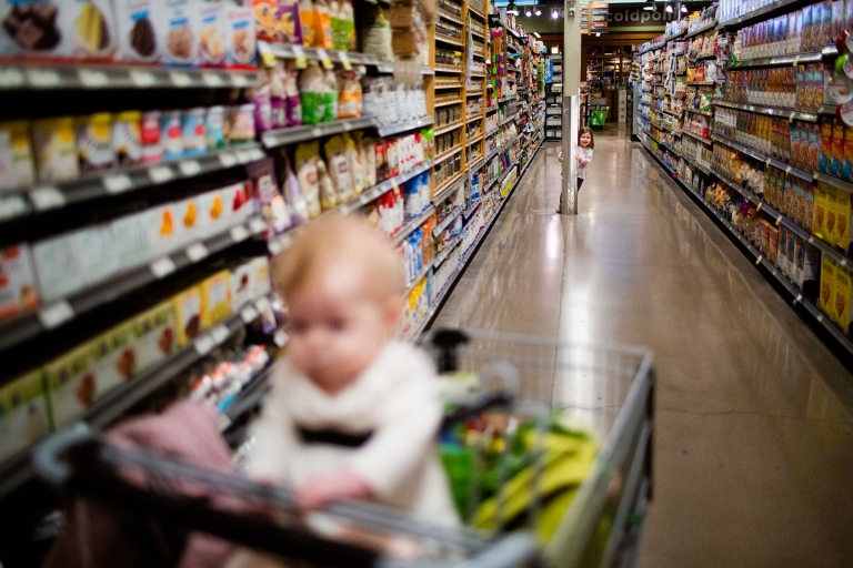 Image in Whole Foods Market of toddler girl peeking out from behind a pole in the background with baby girl sitting on a cart in the foreground.