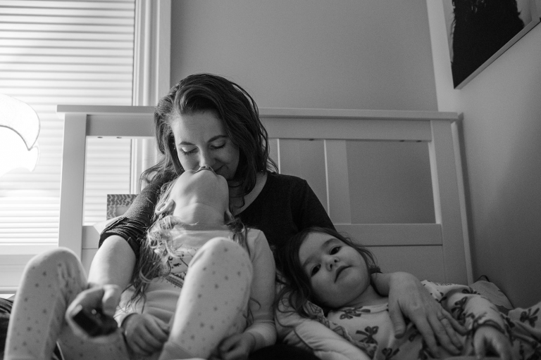 black and white image of mom on a bed with two little girls, one looking at camera and the other kissing her