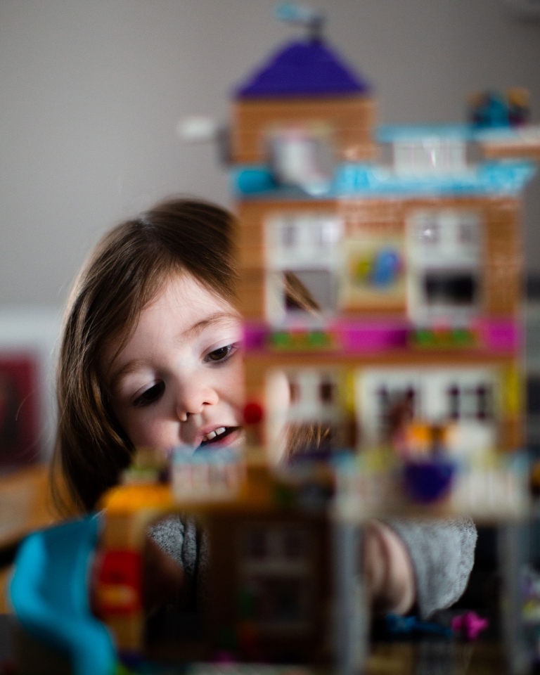 Photograph of little girl playing with legos with a lego house in the foreground.