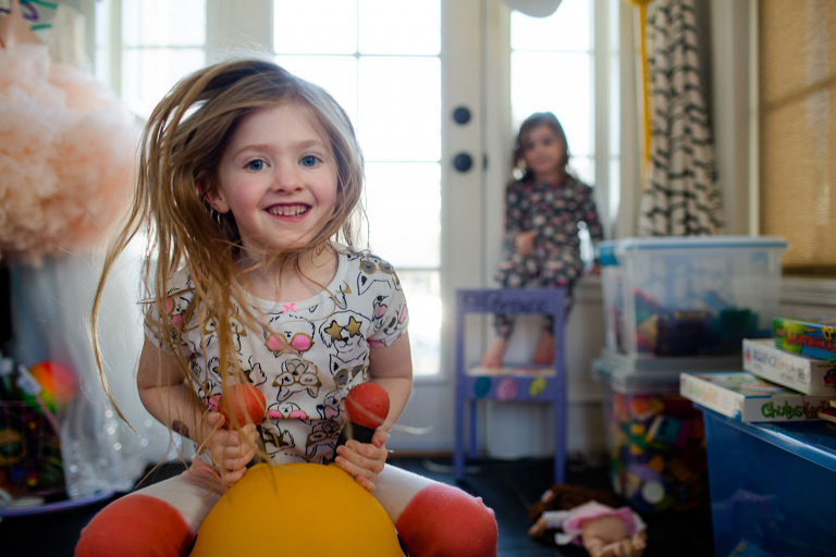 Little girl bouncing on bee toy with toddler little sister looking on in the background in messy playroom