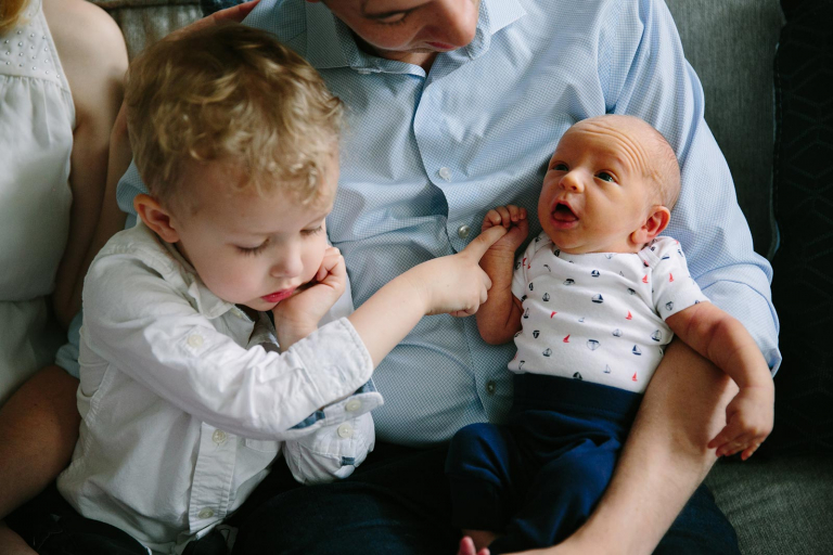 Documentary Photograph of little boy and newborn boy sitting on dad's lap holding hands