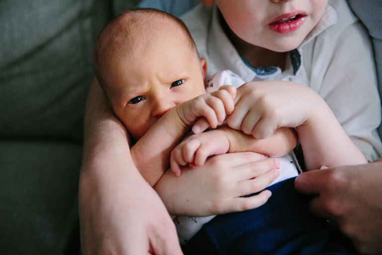 Closeup of newborn sitting on big brother's lap, holding his finger and glaring at camera