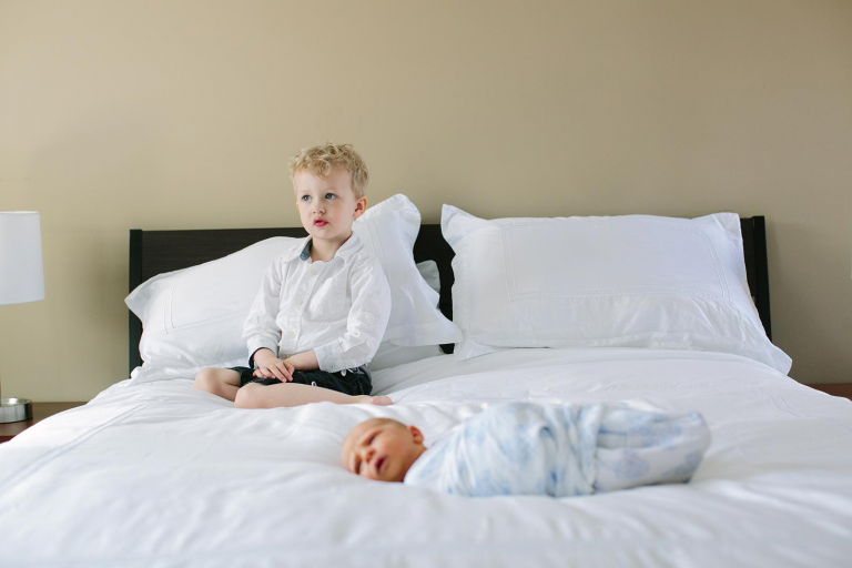 Photograph of newborn swaddled on a bed with big brother sitting on the bed in the background