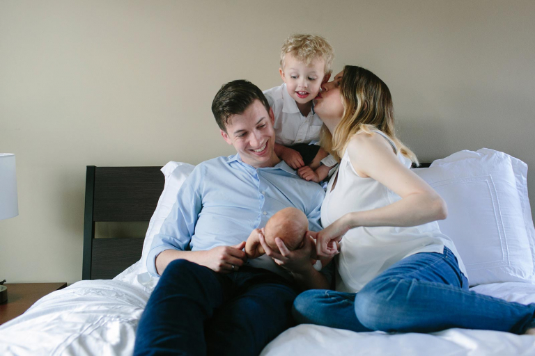 Newborn image of big brother peeking over mom and dad's shoulder and looking at new brother