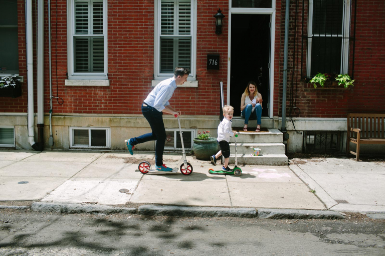 Documentary photograph of mom sitting on front step with newborn and dad and little boy riding scooters on the sidewalk